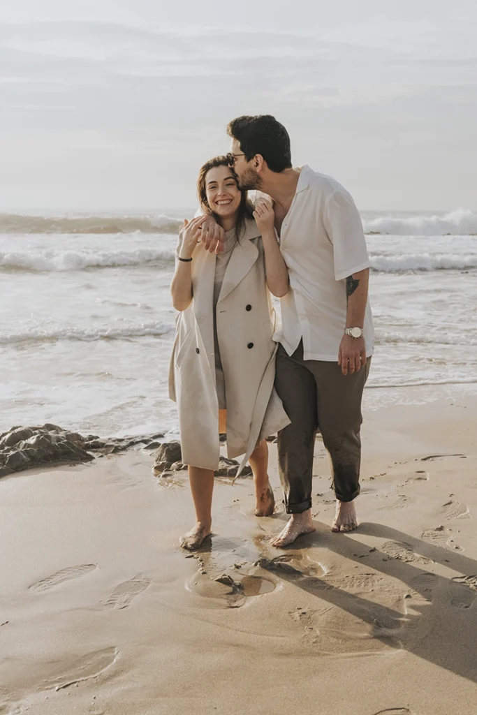 Casal caminhando de mãos dadas na praia, se beijando enquanto desfrutam de um momento romântico à beira-mar.