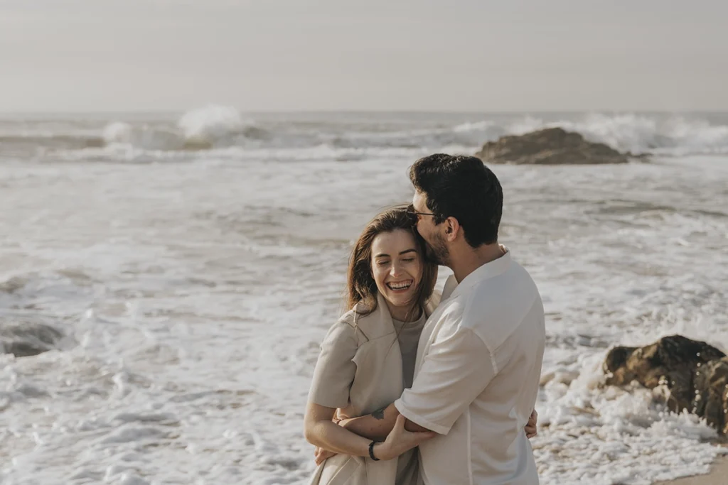 Casal abraçado e sorrindo na praia, com ondas suaves ao fundo, refletindo um momento de felicidade e conexão.