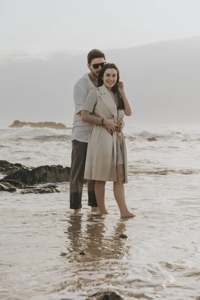 Casal sorri abraçado no mar gelado, celebrando o amor apesar do frio.
