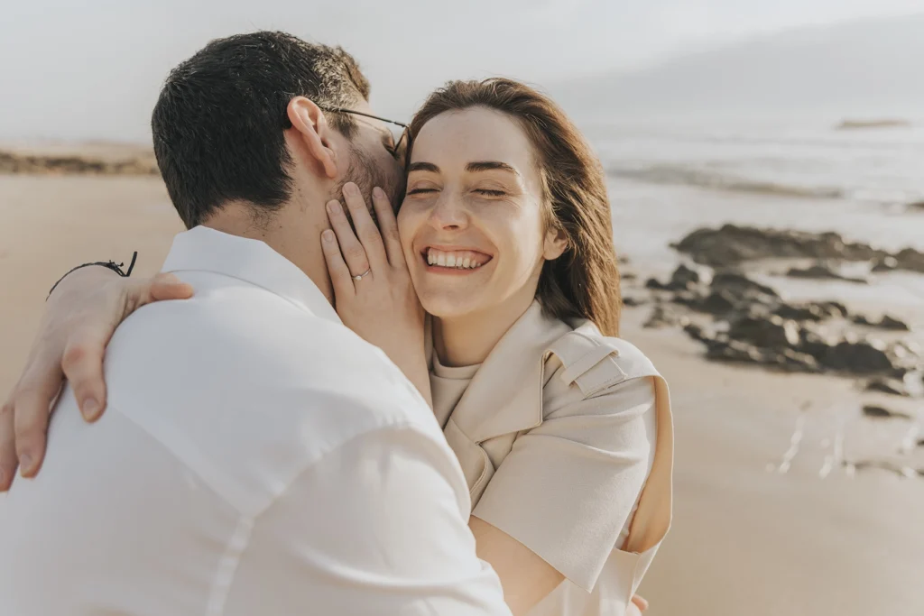 casal abraçado e feliz, sorri mostrando o anel de noivado na praia