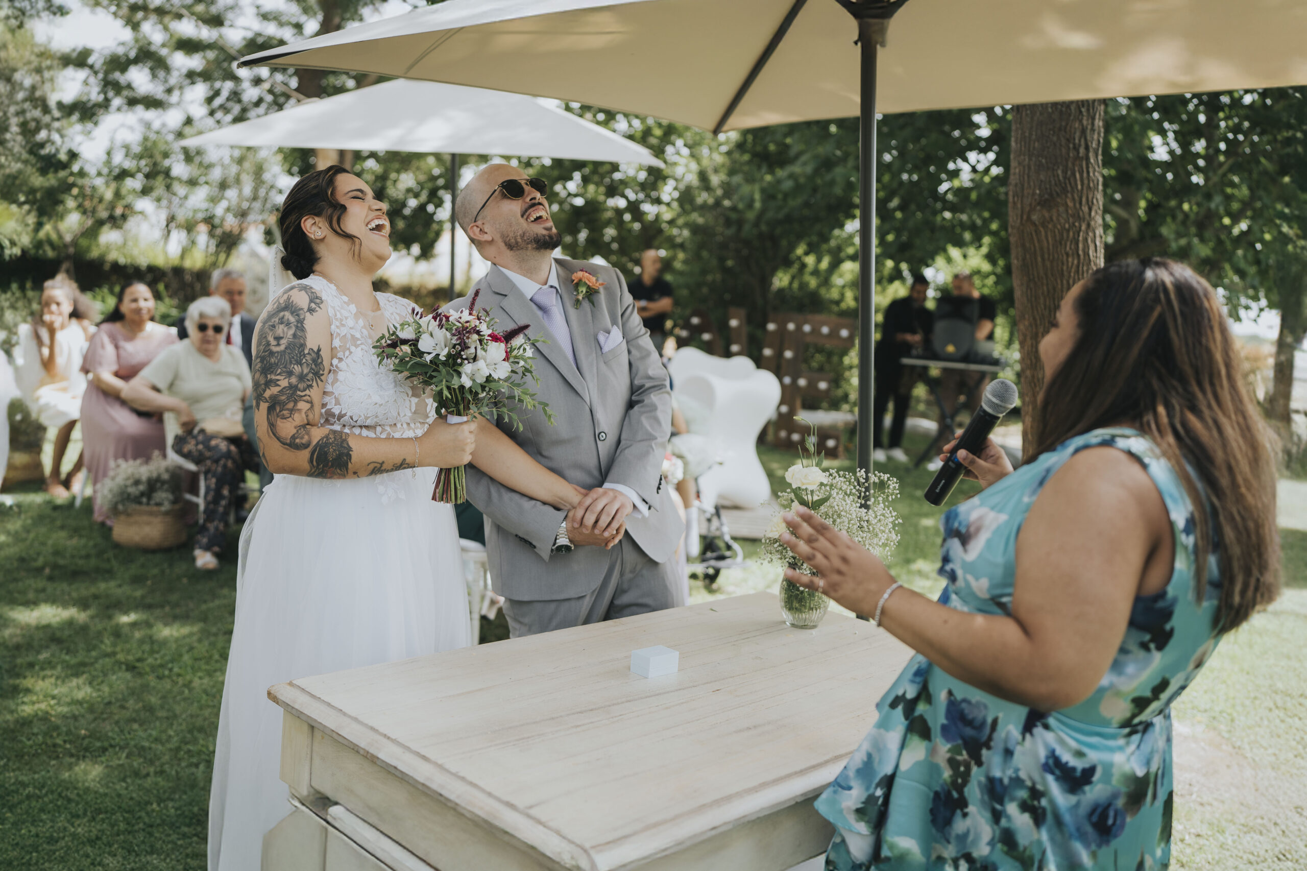 Marah e Miguel, casal de noivos, a rir juntos durante a cerimônia de casamento na Quinta do Nevada, Palmela, rodeados por convidados e vinhedos.